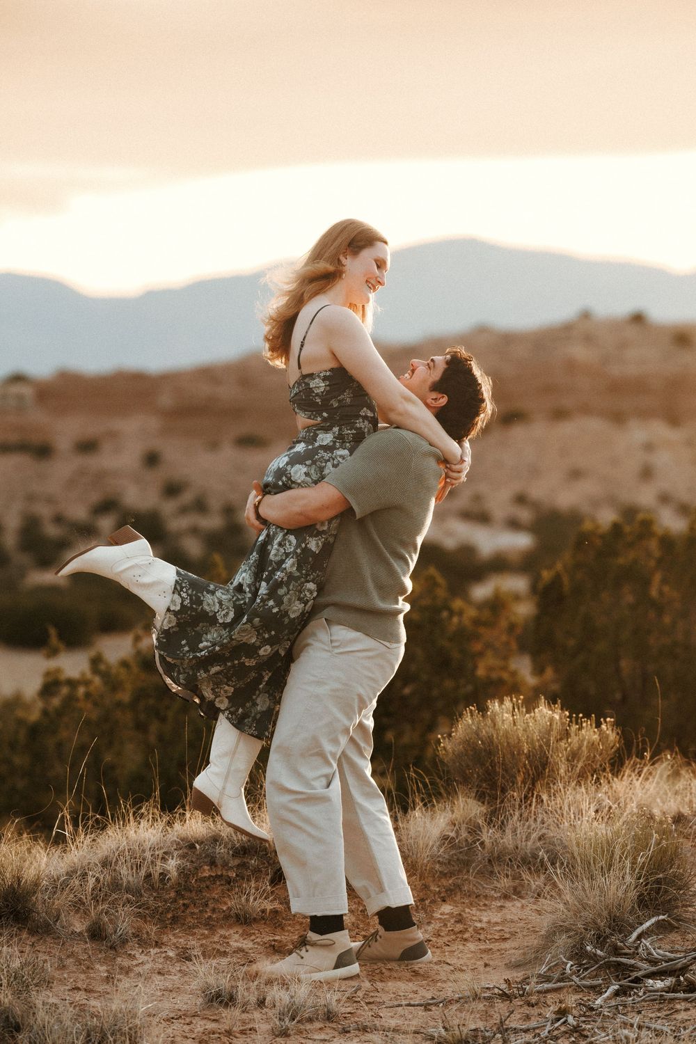 Man lifting woman in floral dress during sunset engagement shoot in scenic desert landscape with mountains.