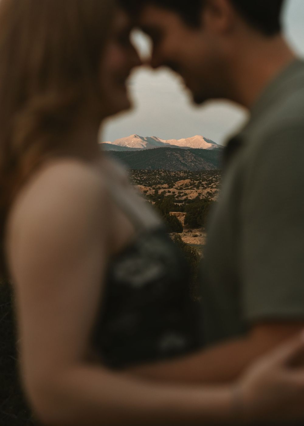 Blurred couple nearly kissing with snow-capped mountains and desert landscape in sharp focus behind them.