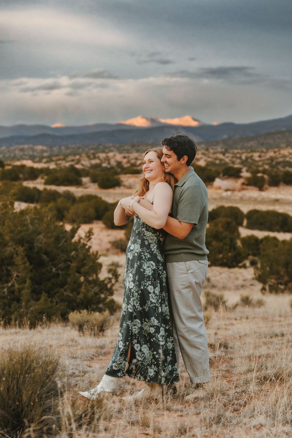 Couple embracing and smiling in desert landscape at sunset with mountains in background.