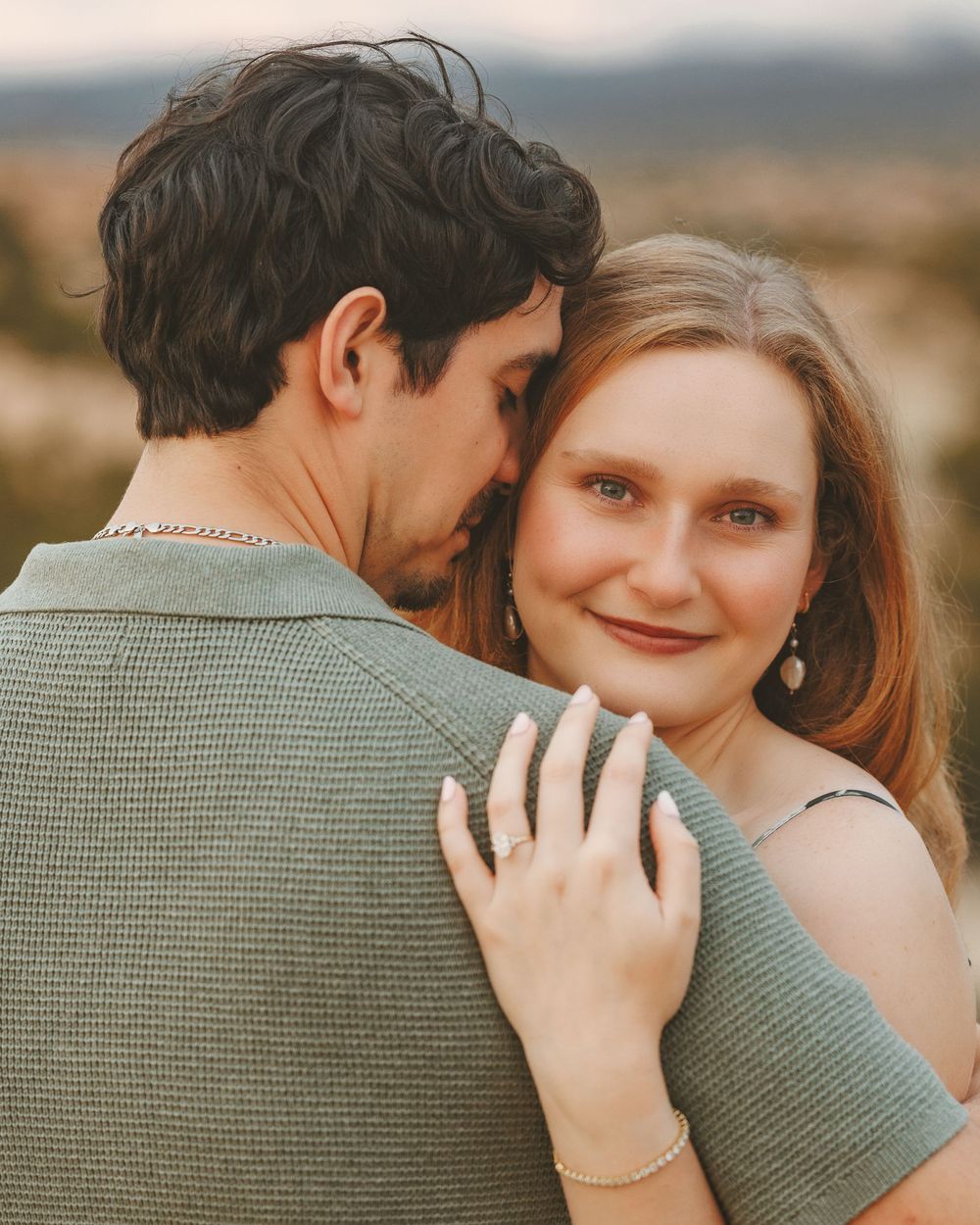 Young woman smiling warmly as her partner embraces her outdoors, showcasing an engagement ring on her hand.