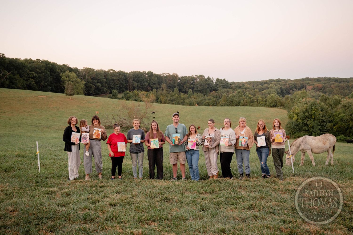 Group of people holding framed photos standing in a green field at dusk with a white horse nearby.