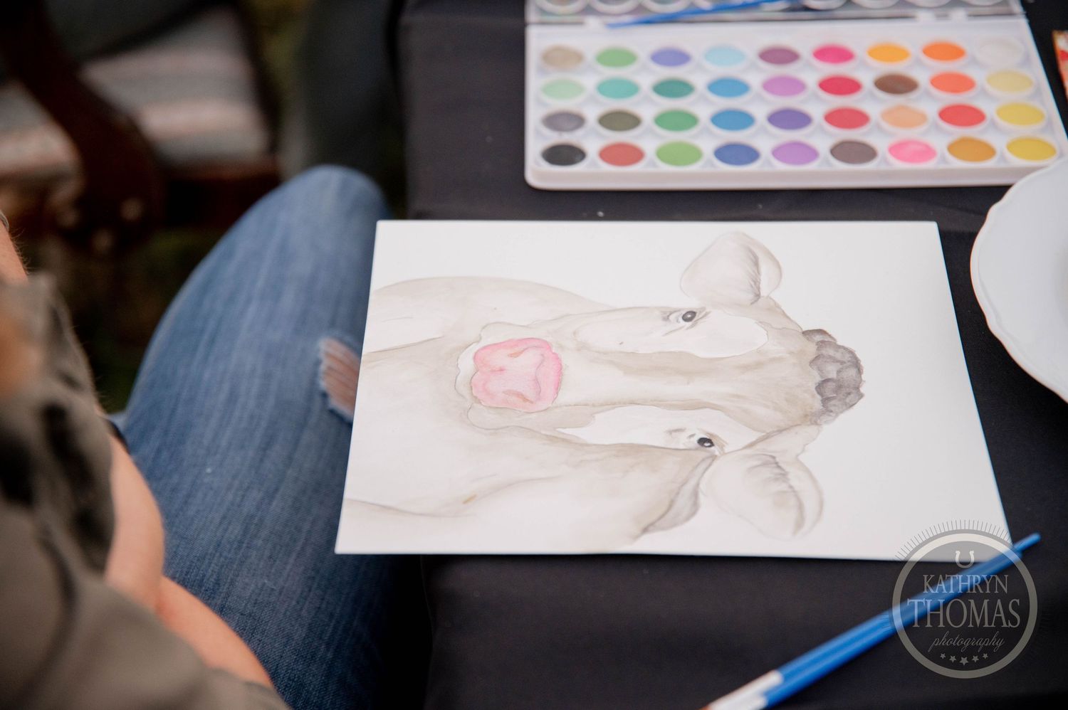 Artist painting a watercolor portrait of a dog on white paper with a colorful paint palette nearby.
