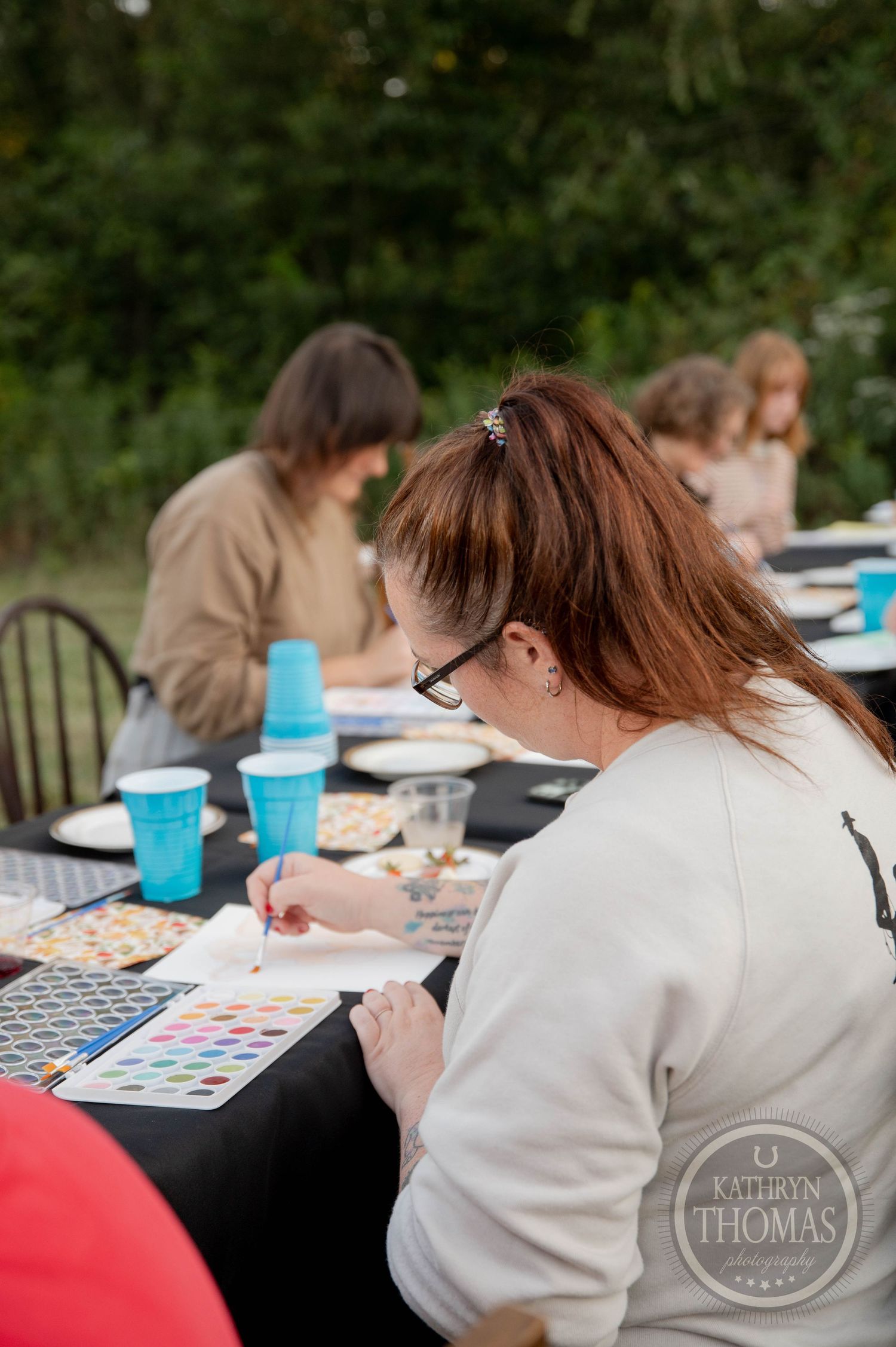 Woman with red hair painting at outdoor art class table with blue cups and other participants in background.