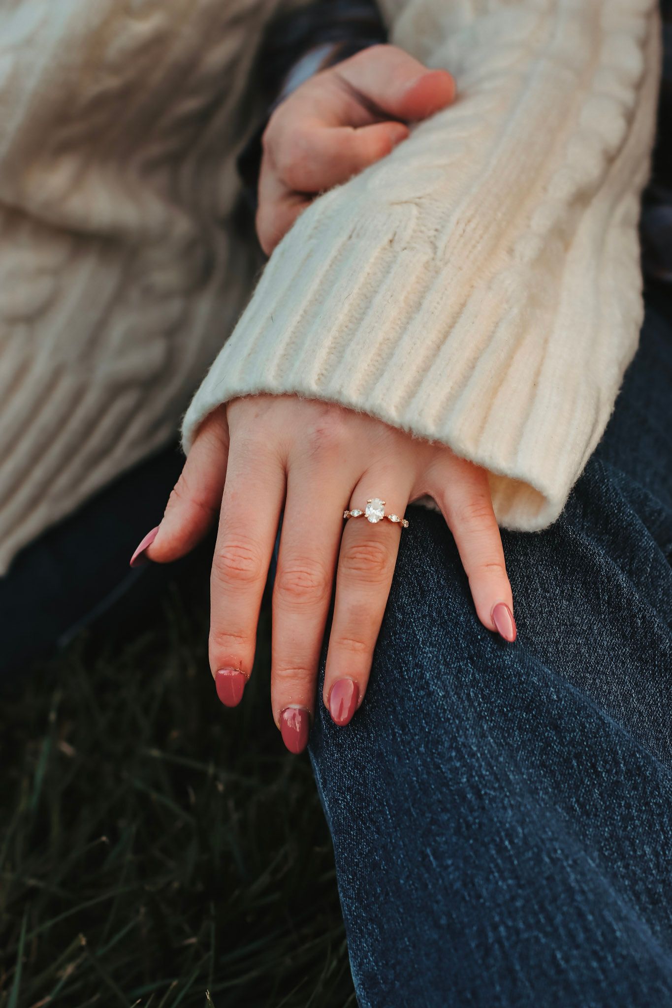 Close-up of a woman's hand wearing a diamond engagement ring, with red nails, resting on denim jeans.