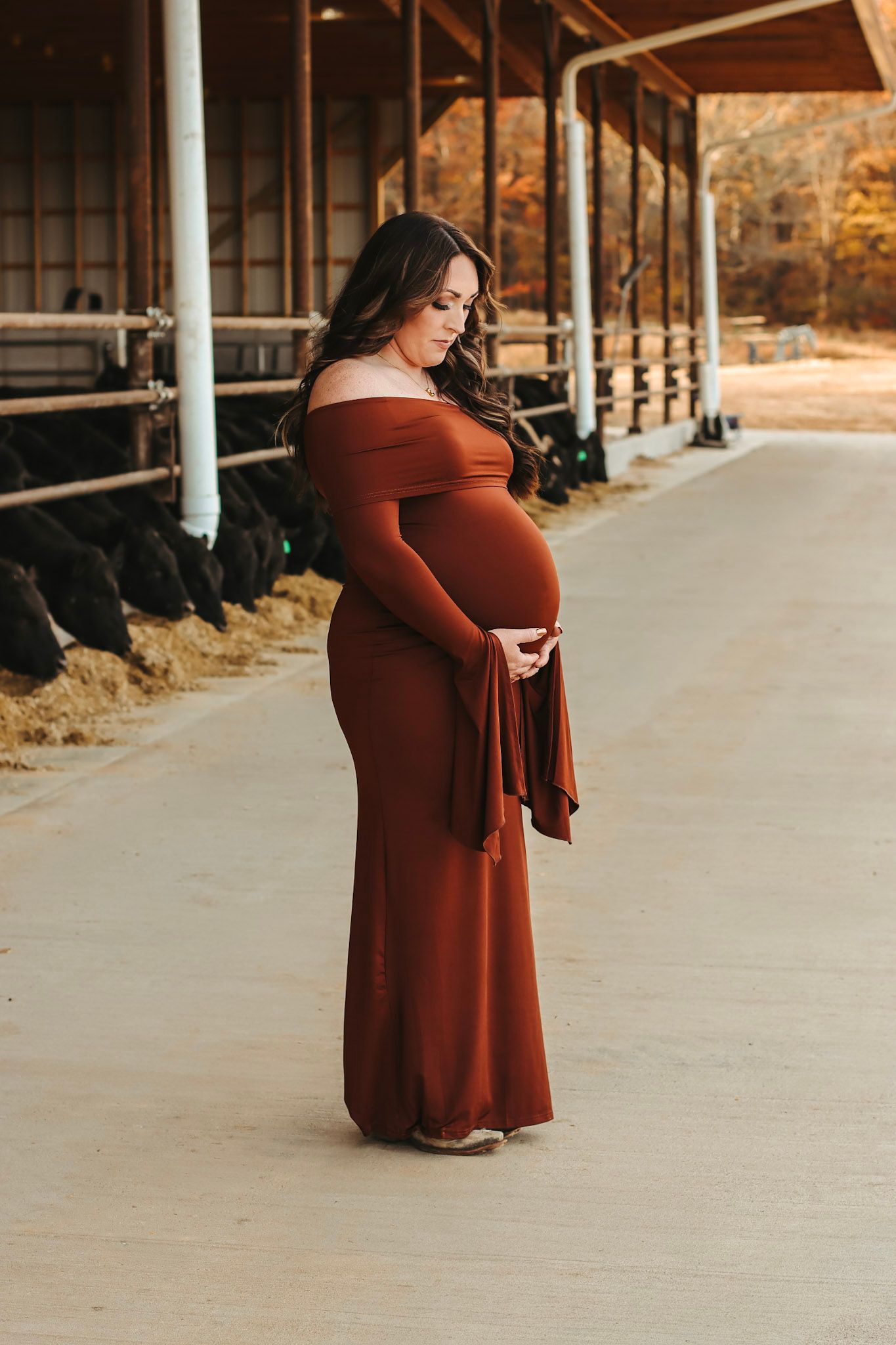 Pregnant woman in rust off-shoulder maxi dress cradling baby bump at a farm barn during maternity photoshoot.