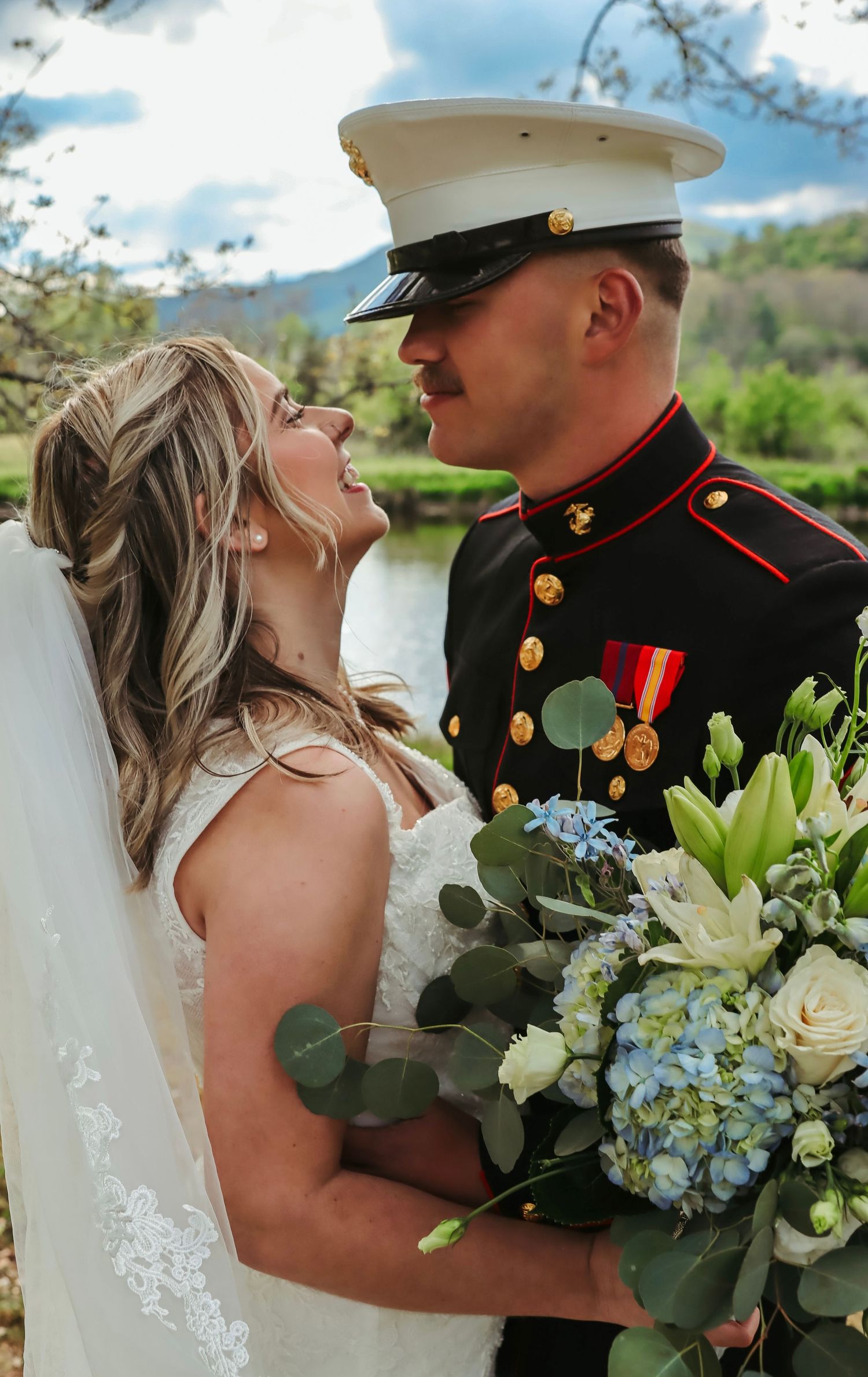 A bride in a white gown holding blue and white flowers gazes up at a Marine in dress uniform outdoors.
