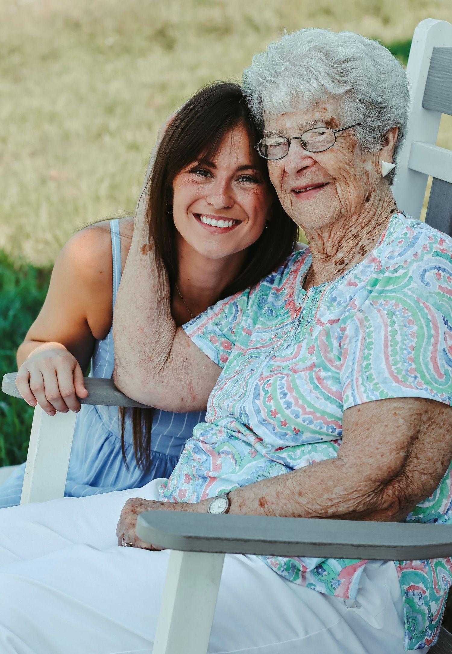 Young woman smiling beside elderly woman with glasses seated outdoors in white chair, sharing a warm moment together.
