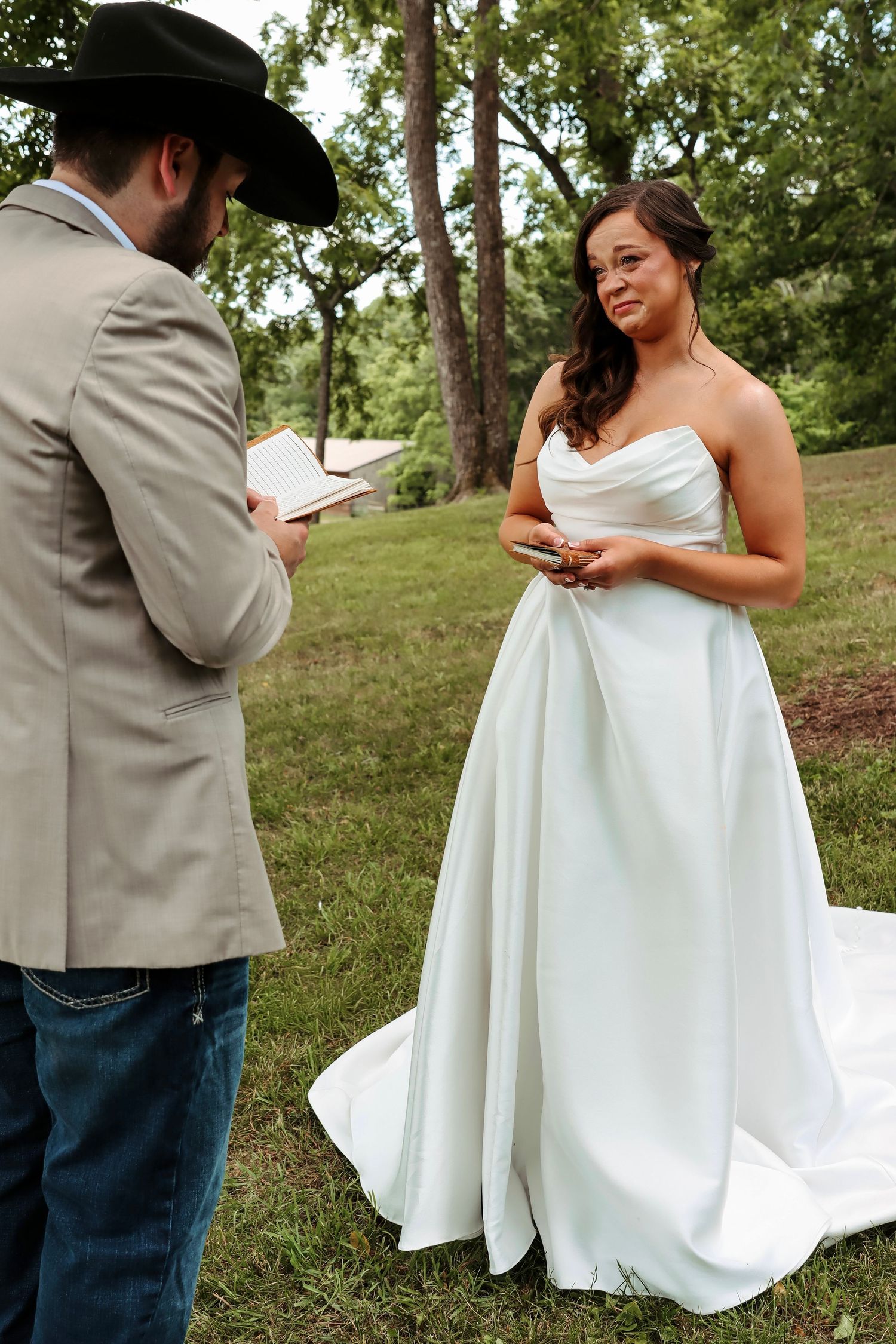 Groom in cowboy hat reads vows to bride in white strapless wedding gown during outdoor ceremony.