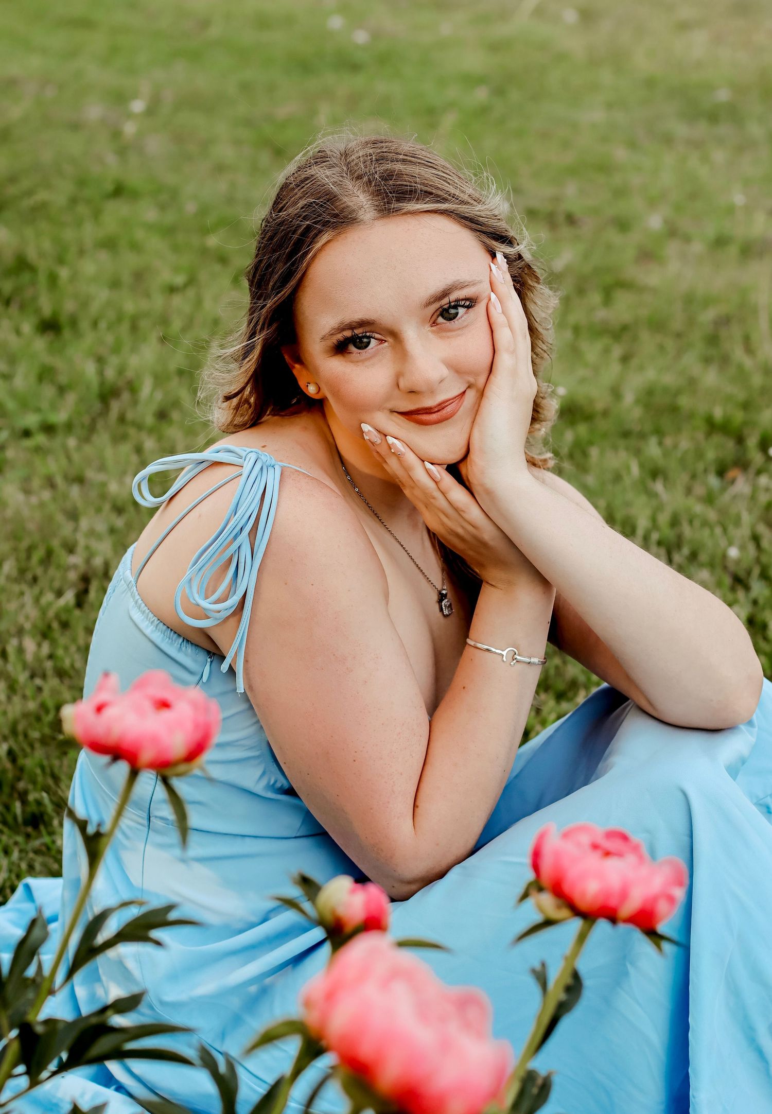 Young woman in blue dress sitting on grass surrounded by pink peonies, smiling gracefully with hand on cheek.