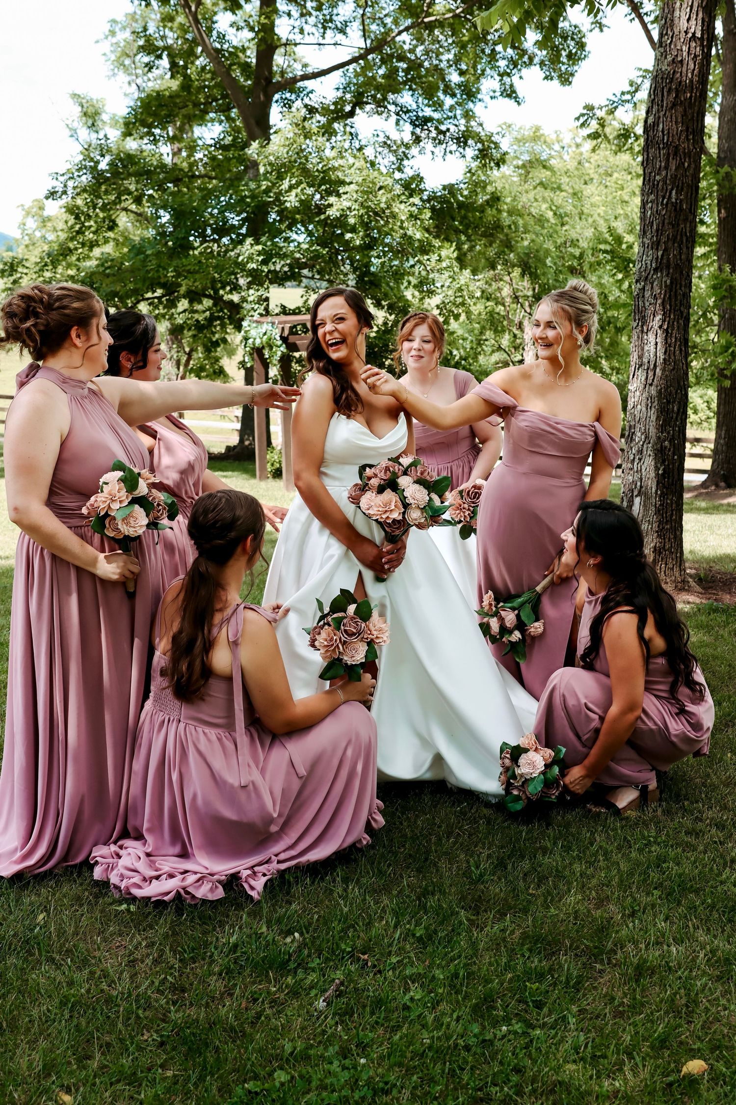 Bride in white gown laughing with bridesmaids in mauve dresses holding pink bouquets outdoors near trees.