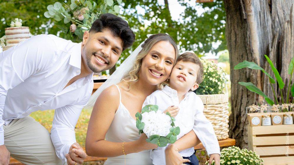 A joyful couple in white wedding attire poses outdoors with their young son, surrounded by lush greenery.