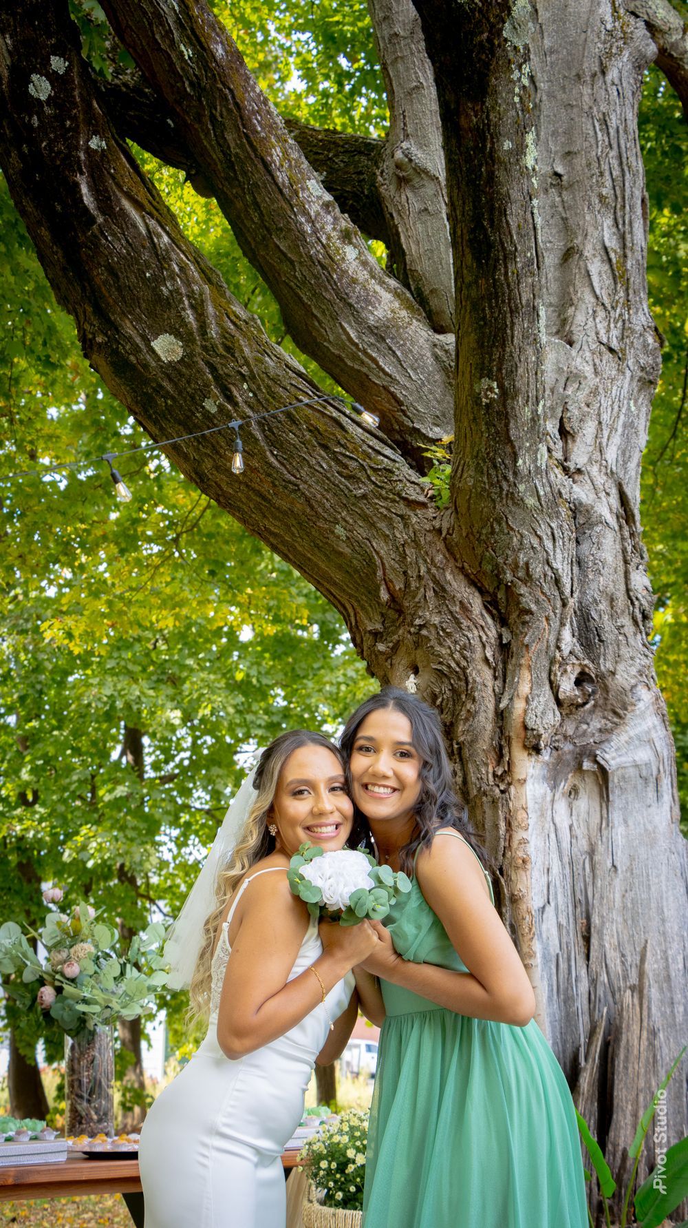 Bride in white gown and veil poses with bridesmaid in green dress under large tree outdoors.