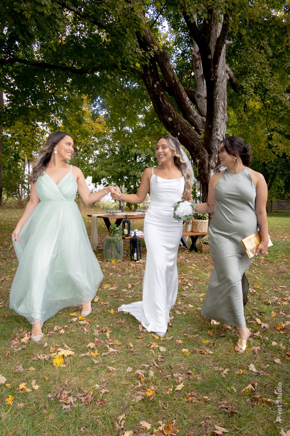 A bride in white holds flowers while walking outdoors with two bridesmaids in sage green and gray dresses.