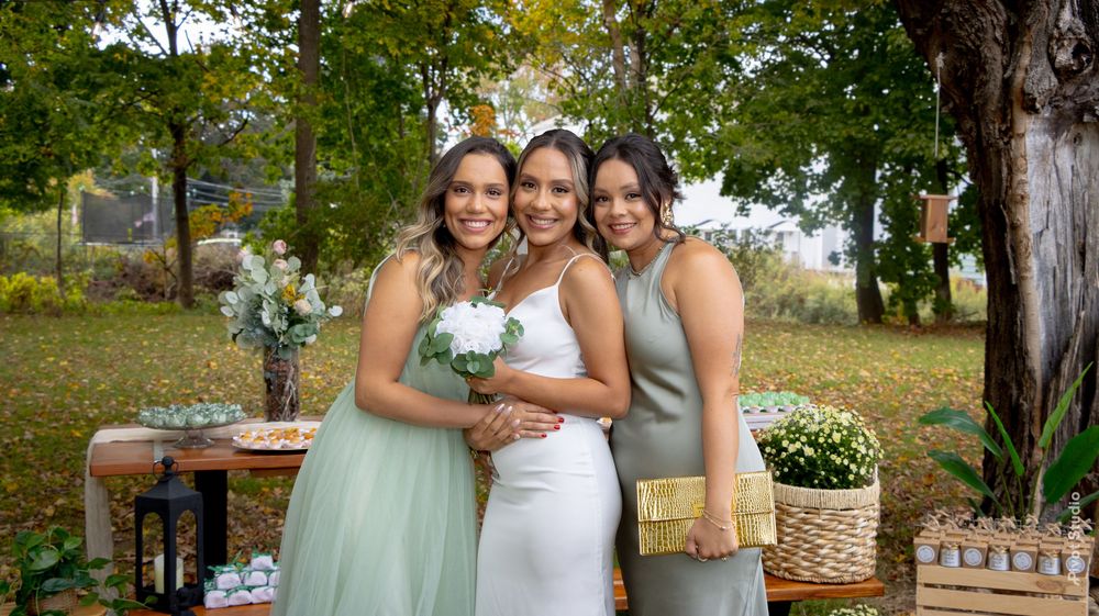 Bride in white gown poses with two bridesmaids in sage green dresses outdoors at a fall wedding reception.