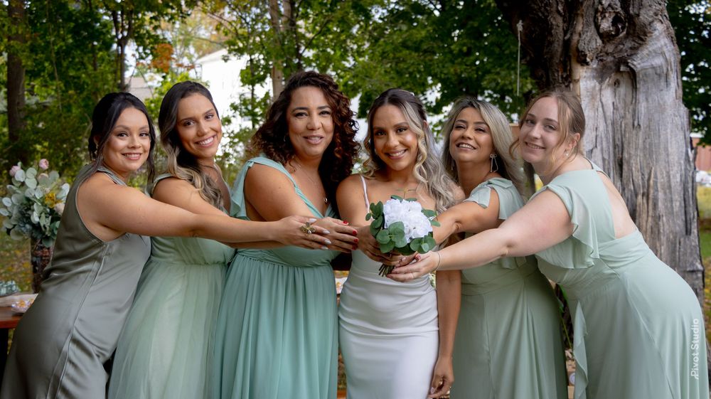 Bride in white gown poses with six bridesmaids in sage green dresses, holding a white bouquet outdoors near a tree.