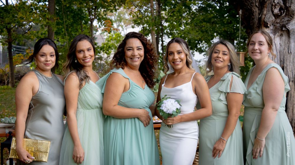 Bride in white dress poses with five bridesmaids in sage green gowns outdoors at a wedding celebration.