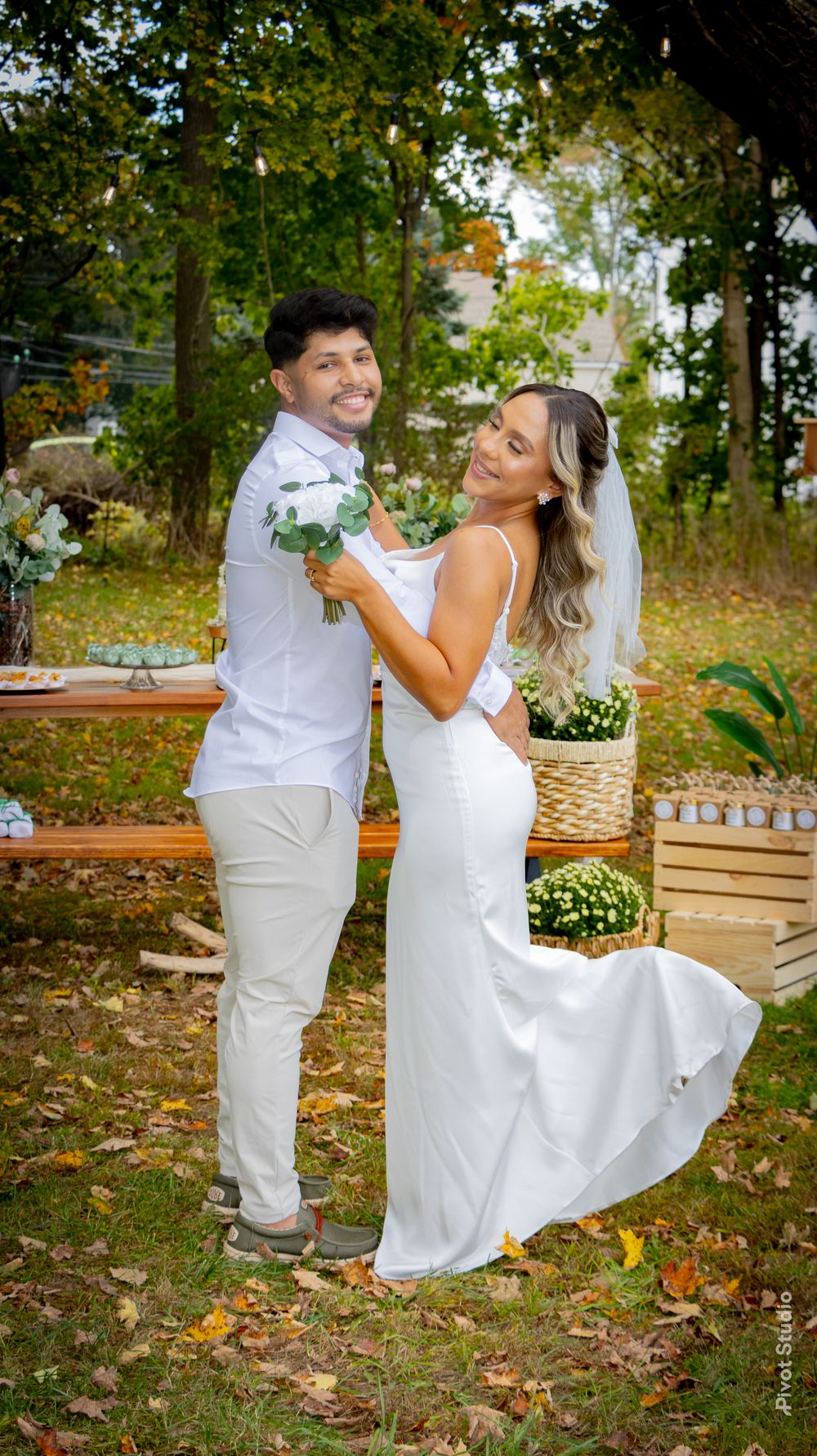 A smiling couple poses outdoors in wedding attire, groom in white shirt and bride in elegant white gown holding a bouquet.