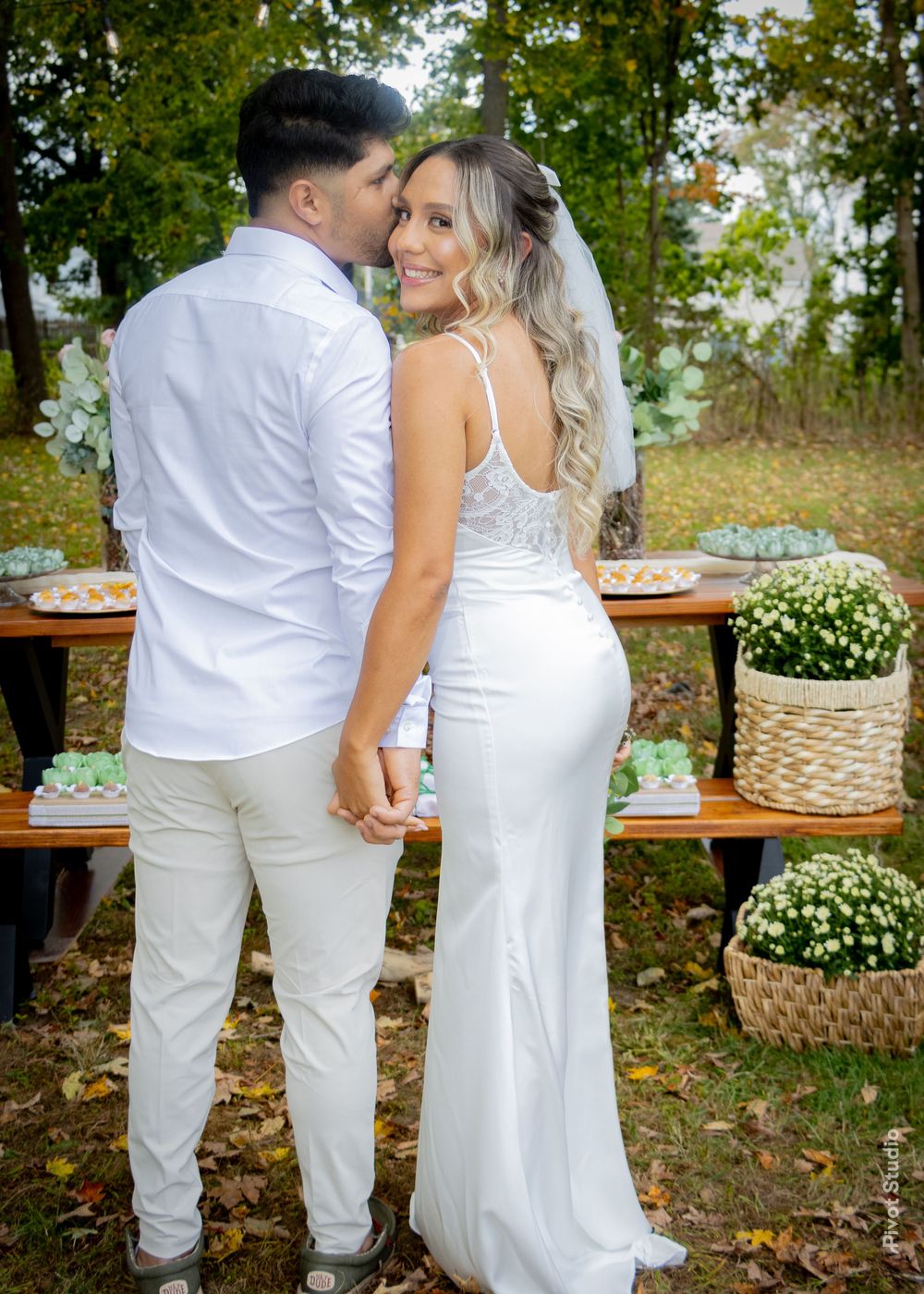 Groom in white shirt kisses bride in wedding gown near decorated outdoor dessert table.