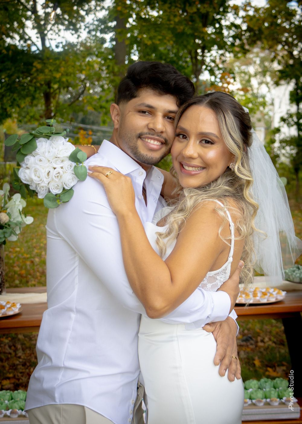 A smiling bride and groom embrace outdoors, wearing white outfits with a floral bouquet at their wedding.