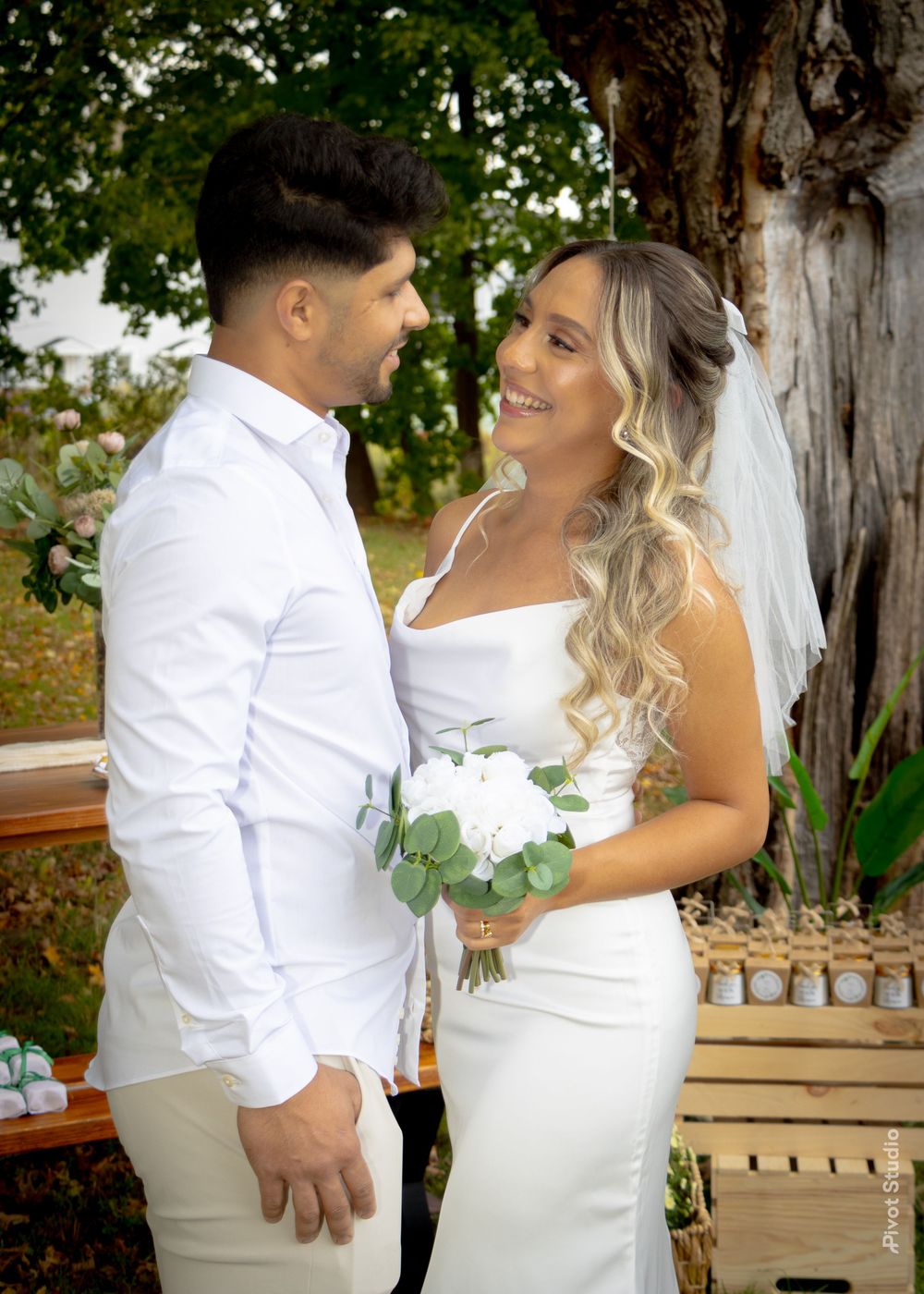Bride and groom in white attire share a romantic moment holding a white bouquet at outdoor wedding.