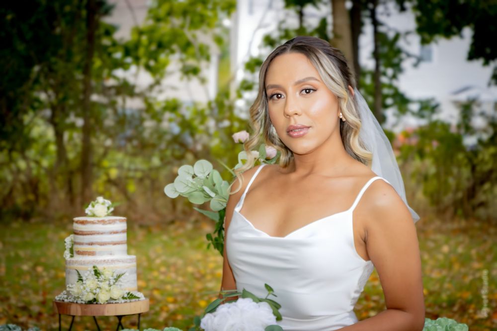 Beautiful bride in white gown with veil poses beside elegant naked wedding cake in garden setting.