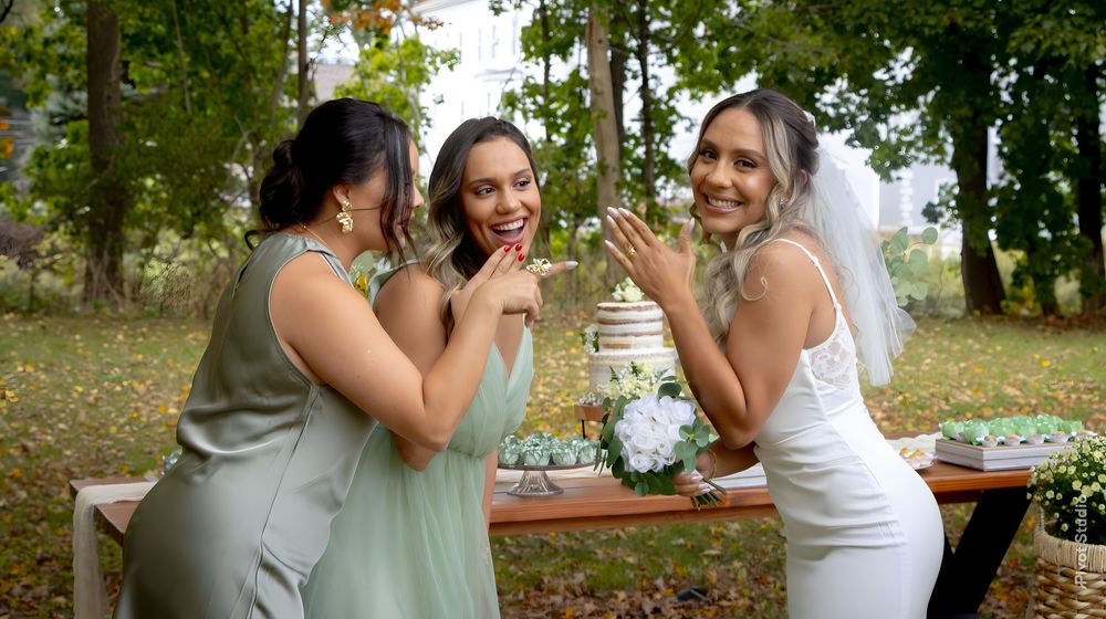 Bride in white gown laughs with two bridesmaids in sage green dresses at outdoor wedding.