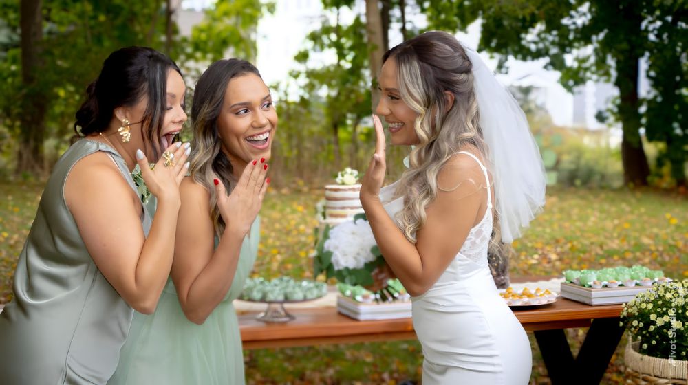 A bride in a white dress and veil laughs with two bridesmaids near a dessert table at an outdoor wedding.
