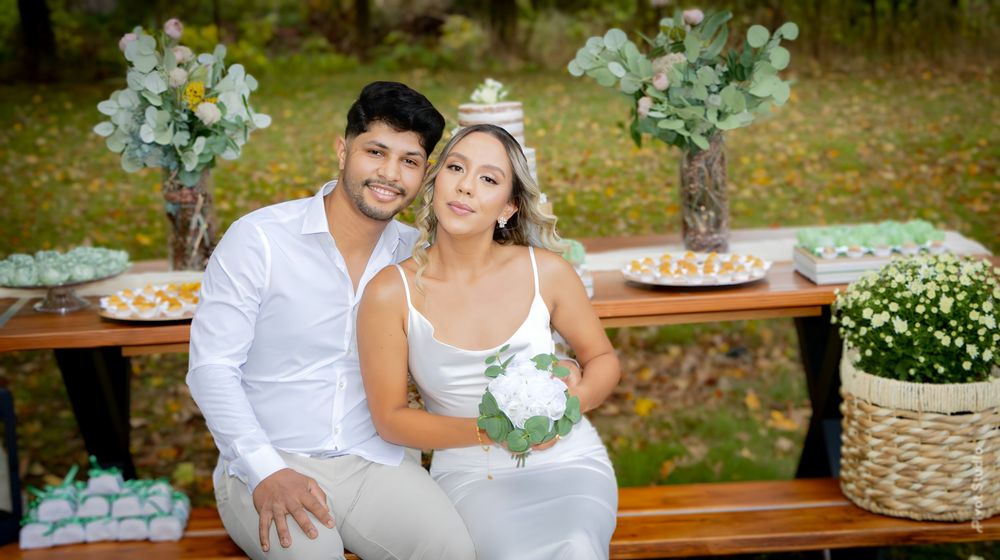 A couple in white attire poses at an outdoor wedding celebration with floral decorations and a dessert table.