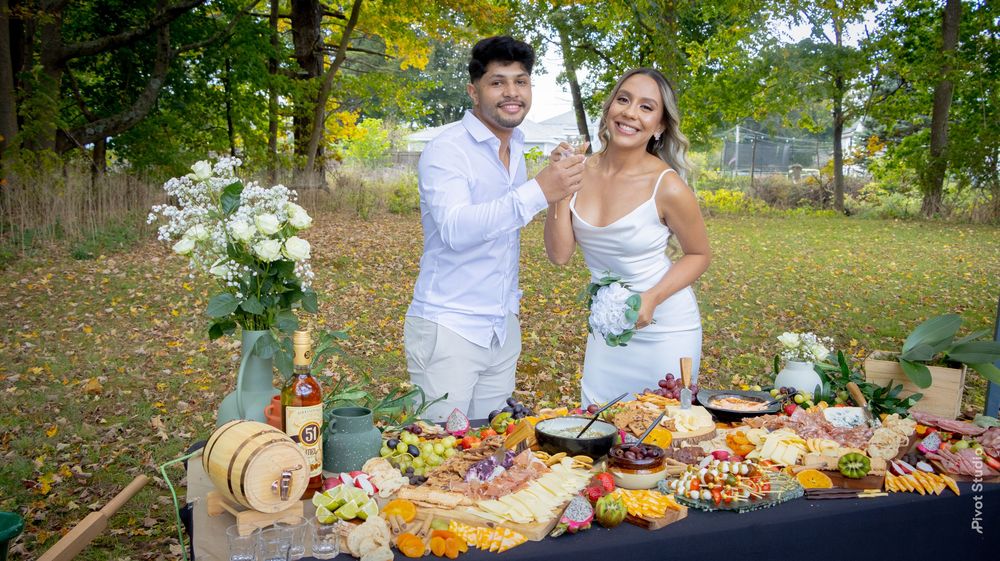 Couple in white outfits smiling at an outdoor picnic charcuterie spread with flowers and fall foliage backdrop.