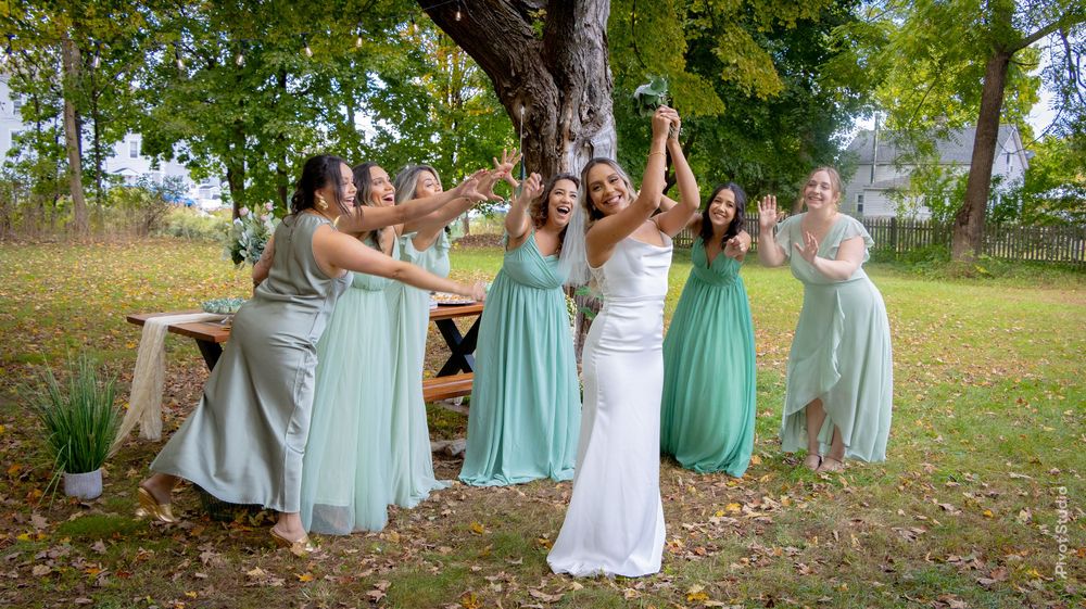 Bride in white gown celebrates with bridesmaids in mint green dresses outdoors near a tree.