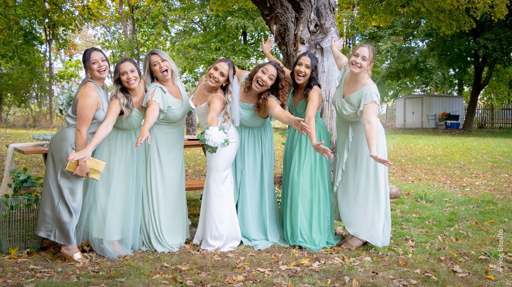 Bride in white gown poses with six bridesmaids in mint green dresses under a tree at an outdoor wedding.