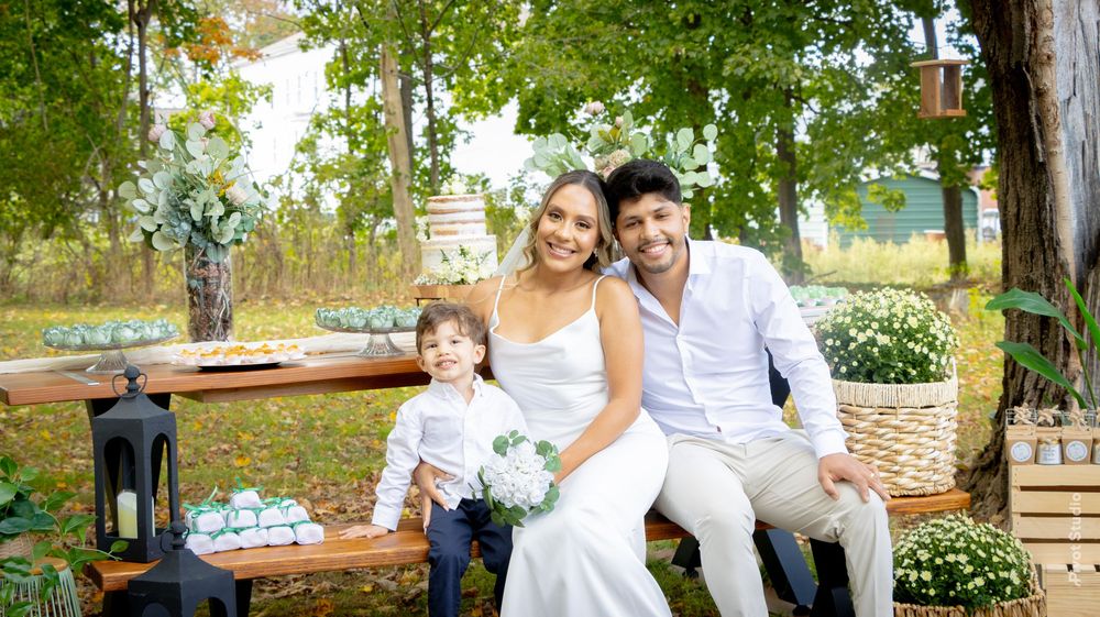 A happy family poses outdoors at a wedding, with a bride in white, groom in cream, and young son in a suit.