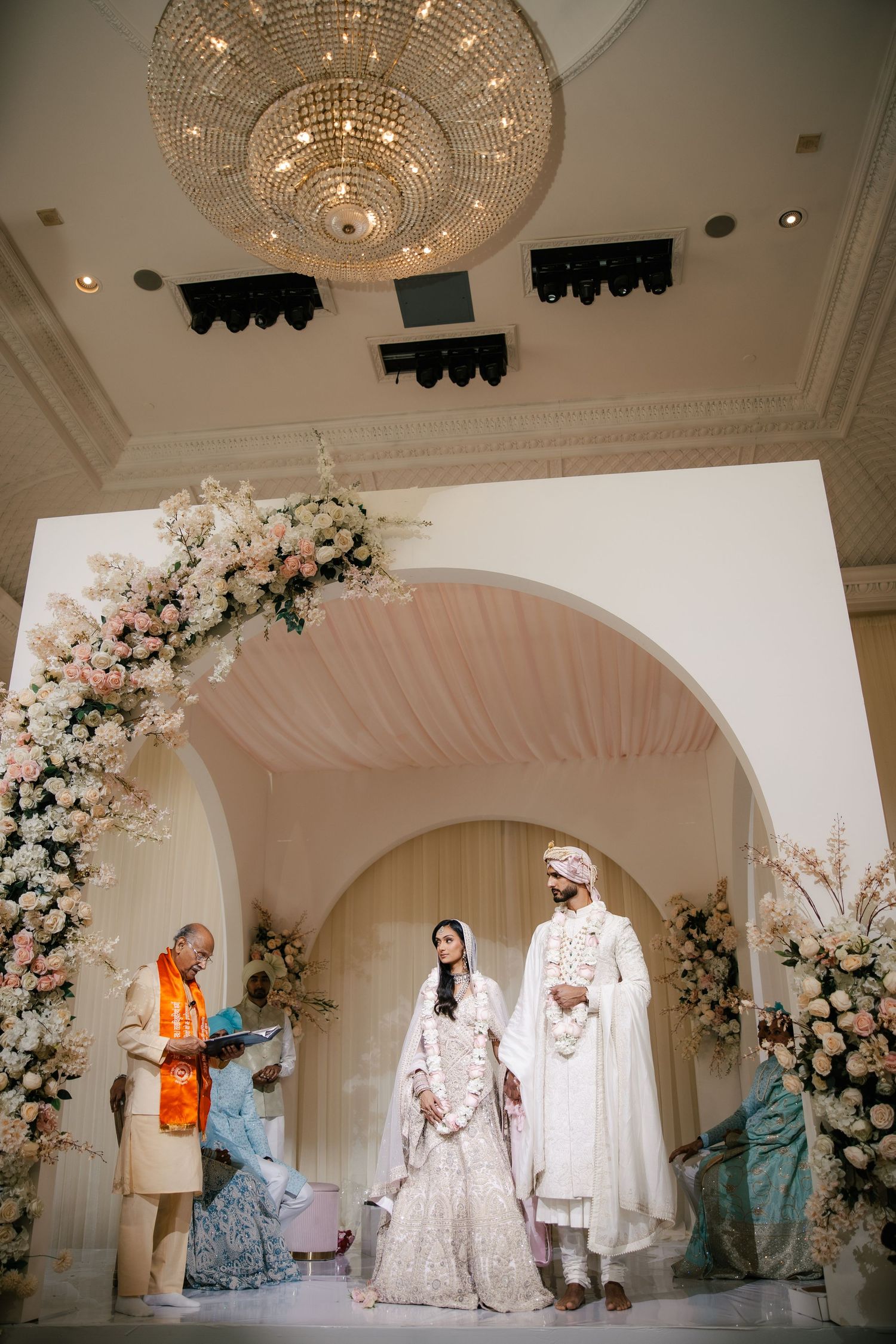 Couple in white wedding attire stand under floral arch at elegant South Asian wedding ceremony with chandelier.