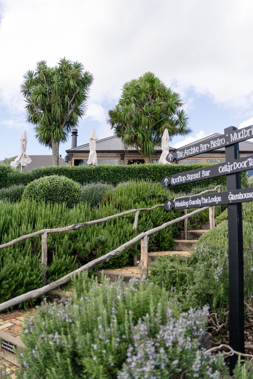 Lush garden pathway with wooden railings, directional signpost, palm trees, and manicured hedges leading to a rustic venue.