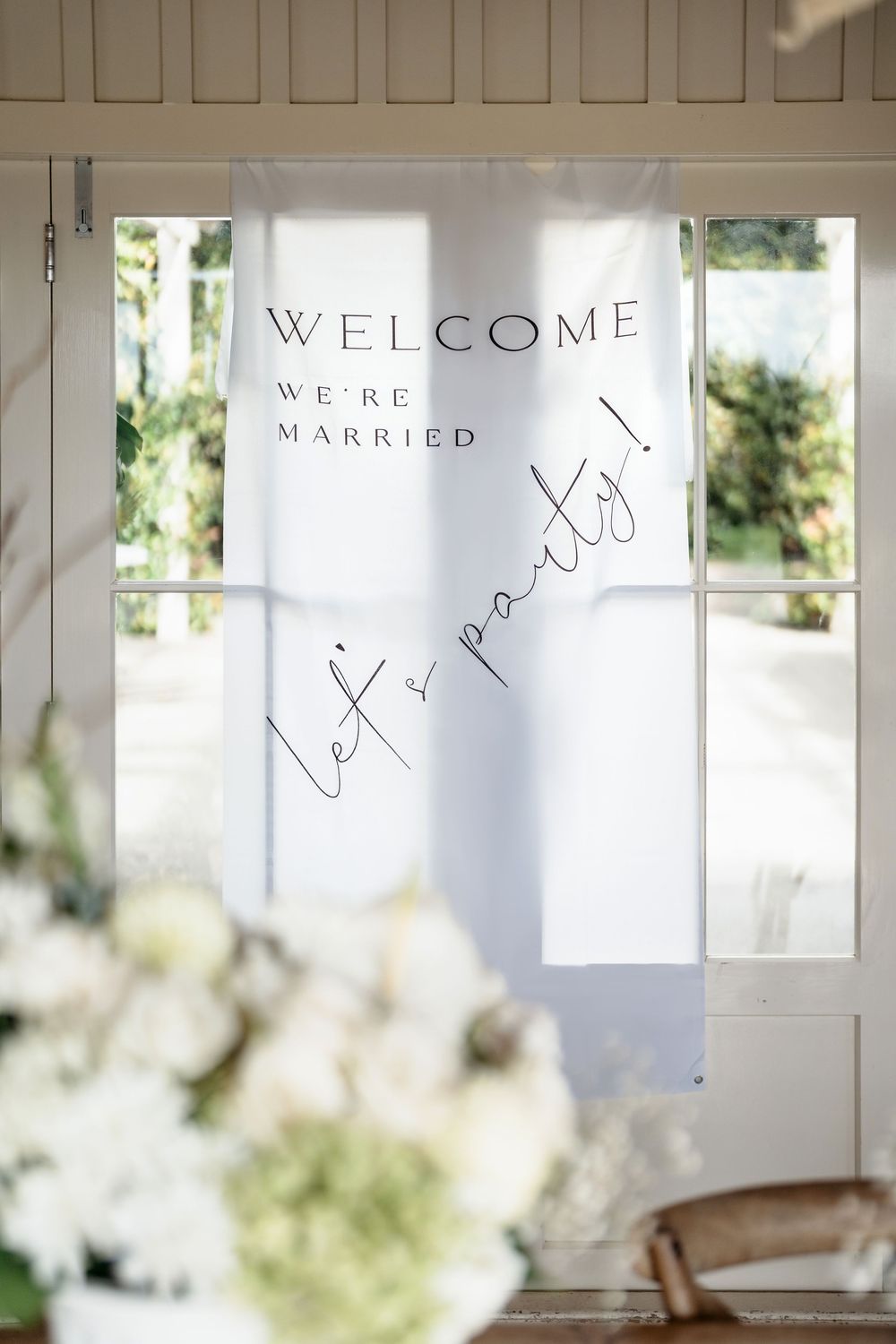 White wedding banner reading Welcome We're Married Let's Party hanging in a bright doorway with floral arrangements.