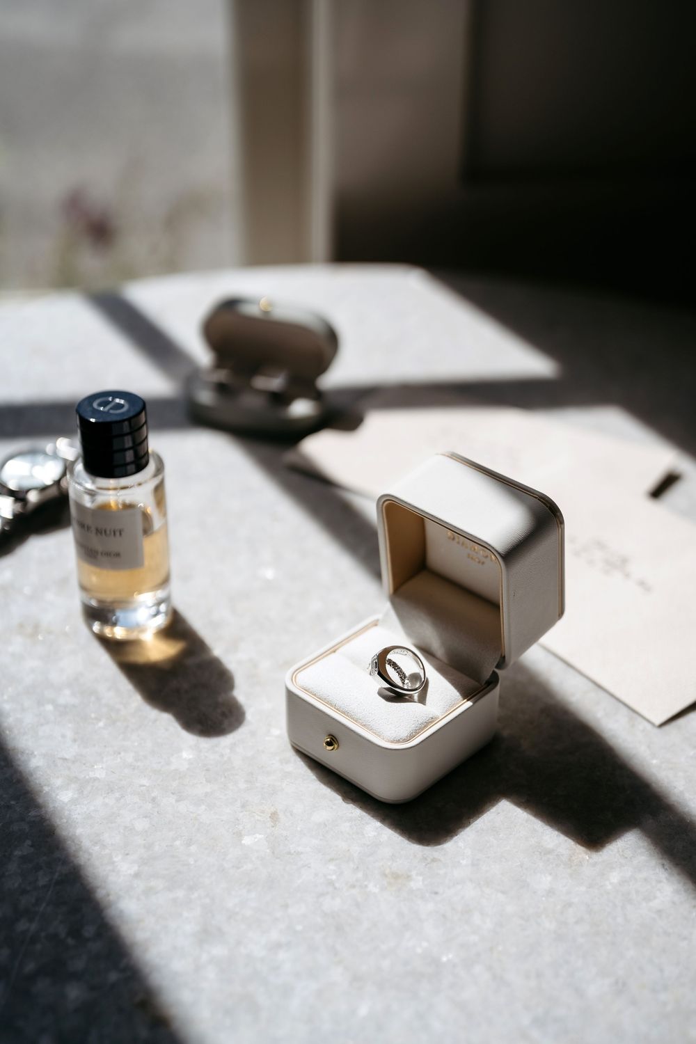 Elegant wedding ring in white box beside Dior perfume bottle on marble surface in natural light.