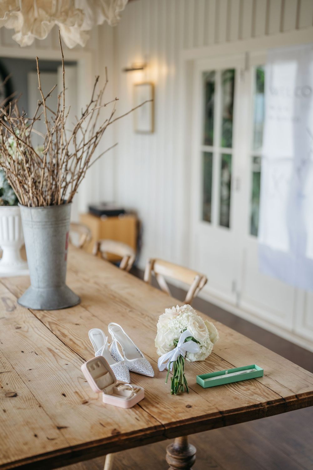 Rustic wooden table with bridal accessories including white heels, ring box, hydrangea bouquet, and a vase of branches.