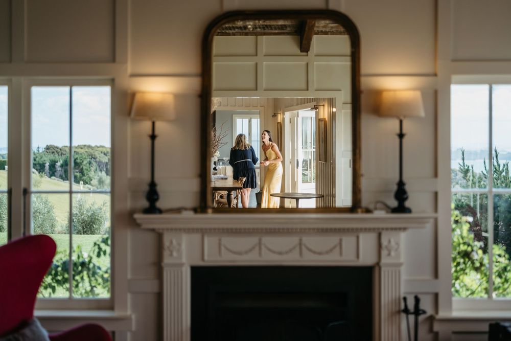Elegant fireplace with ornate mirror reflecting two women in formal attire, framed by large windows with garden views.