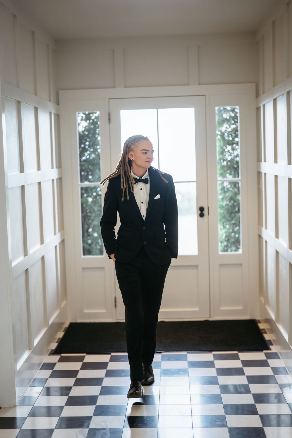 A young woman in a black tuxedo with bow tie walks confidently down a hallway with checkered floors.