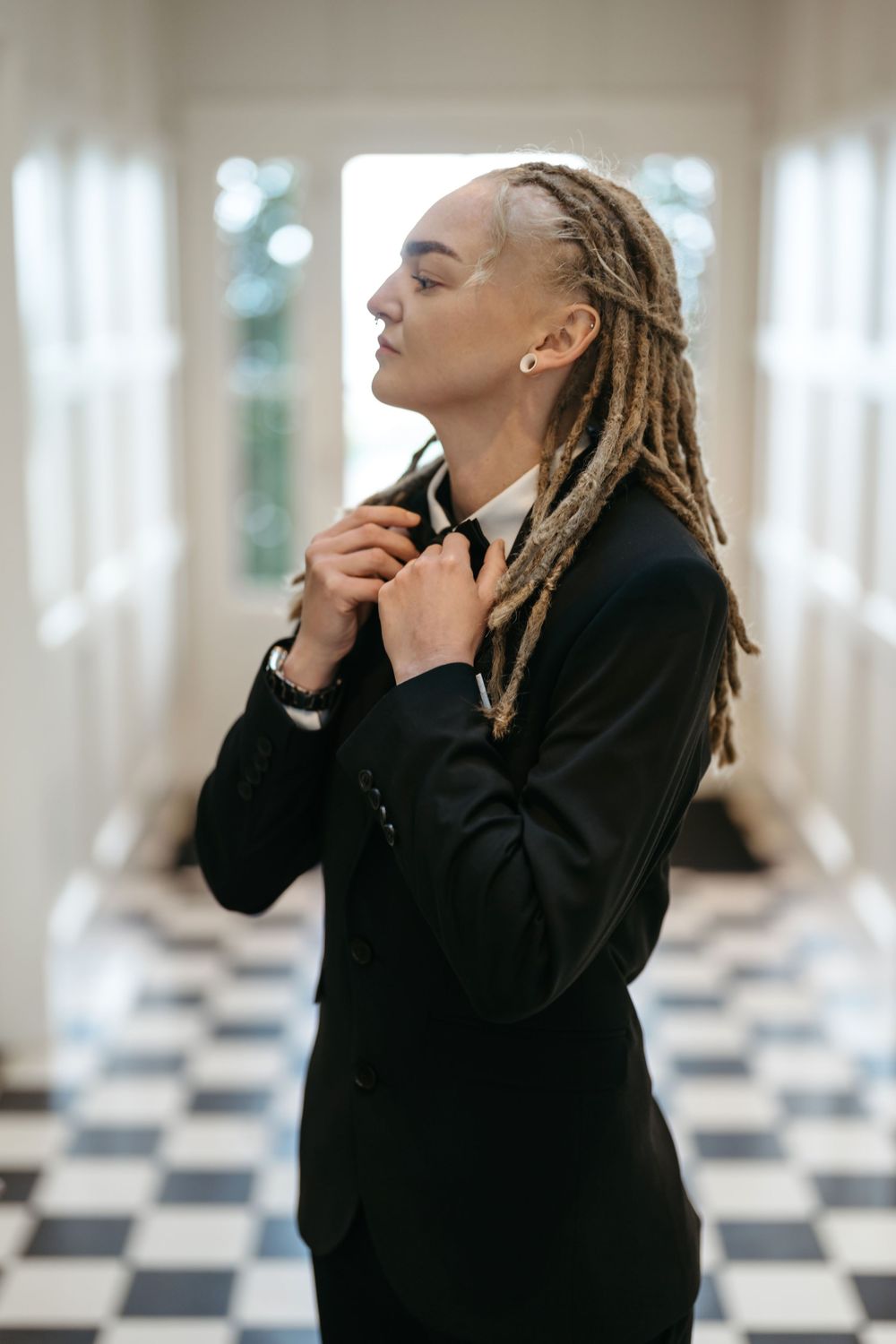 Person with dreadlocks in black suit adjusting bow tie in elegant hallway with checkered floor.