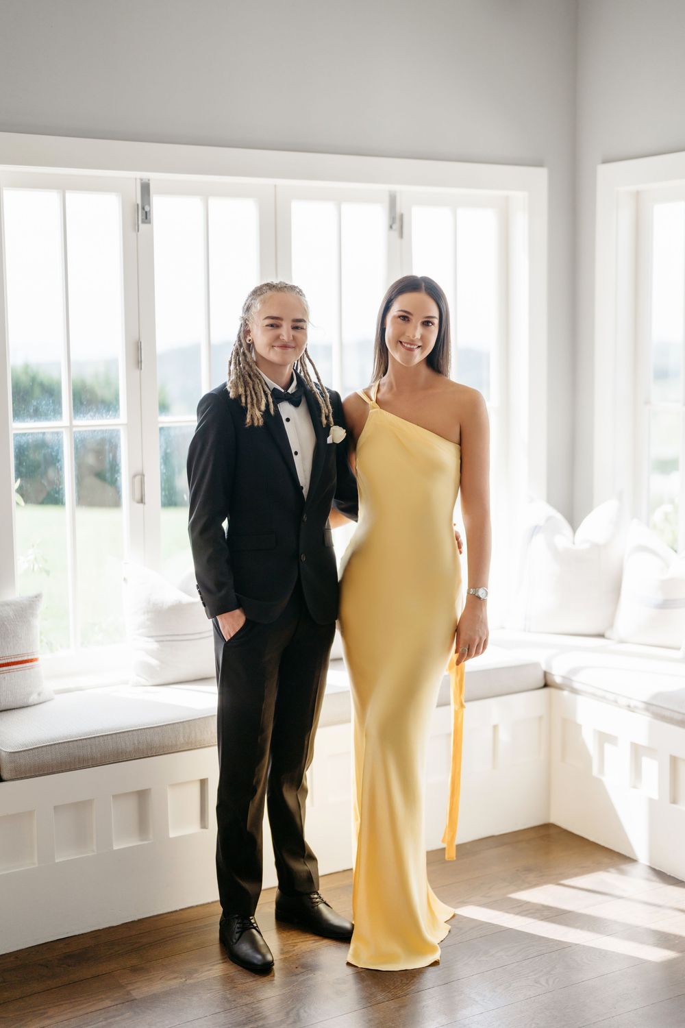 Two teens dressed for prom, one in a black tuxedo and one in a yellow one-shoulder gown, posing indoors.