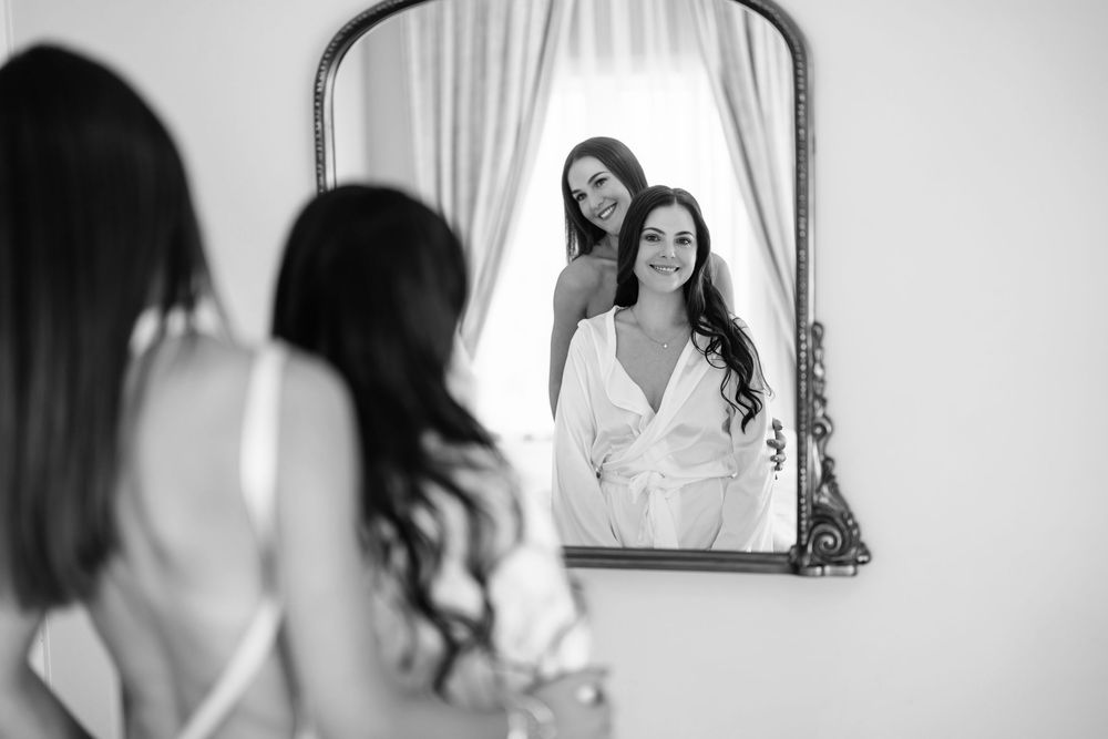 Bride and bridesmaid share a joyful moment reflected in an ornate mirror during wedding preparations.
