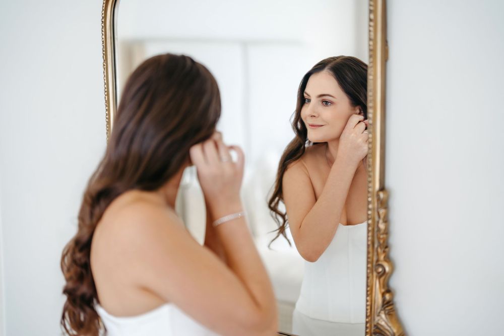 Bride in white dress putting on earrings while looking in an ornate gold mirror before her wedding.