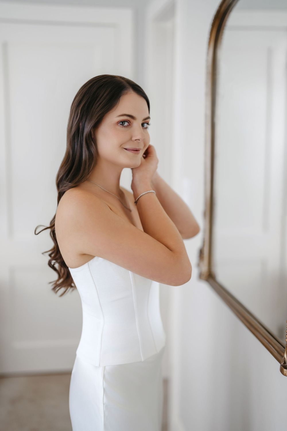 A bride in a white strapless gown adjusts her earring while standing beside a gold-framed mirror.