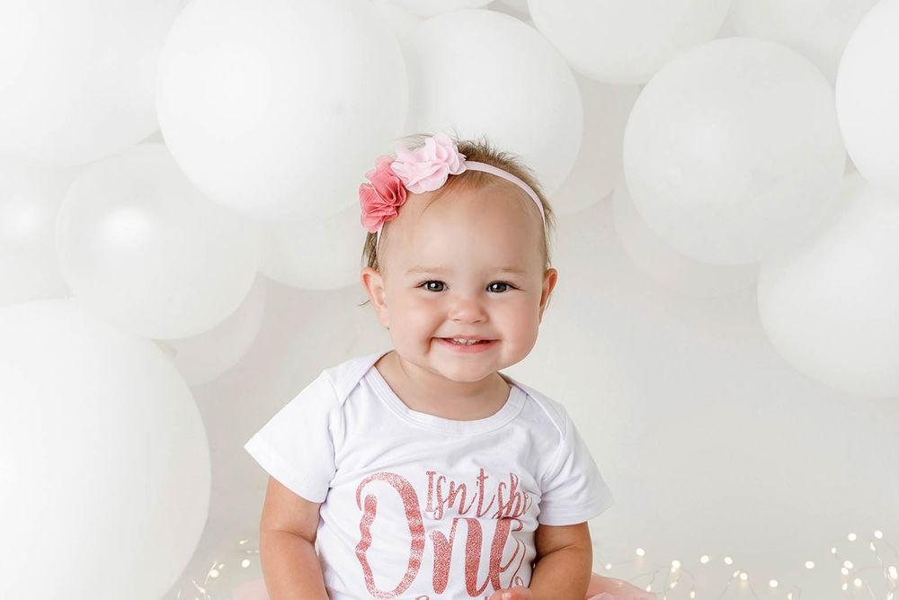 Smiling baby girl in a white “Isn’t She One” onesie and pink floral headband poses in front of white balloons.