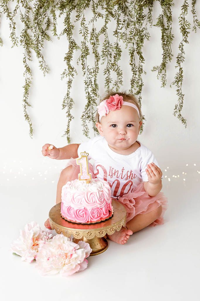 Baby girl smashes pink rosette first birthday cake on gold stand in white studio setting by Temescal Valley cake smash photographer.