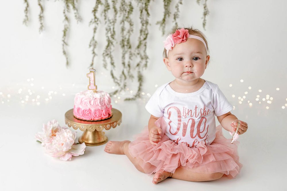 Adorable baby girl in pink tutu sits beside first birthday cake with floral headband.