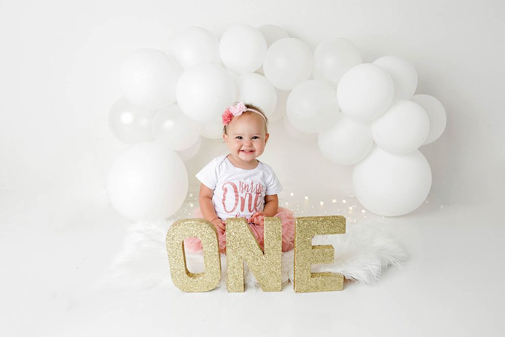 Baby girl sits in front of gold ONE letters and white balloon arch for first birthday by Temescal Valley cake smash photographer.