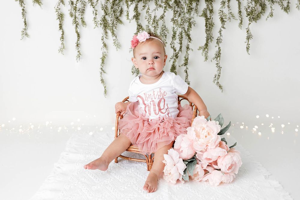 Baby girl in pink tutu dress sits in wicker chair with floral backdrop for first birthday photo by Temescal Valley cake smash photographer.
