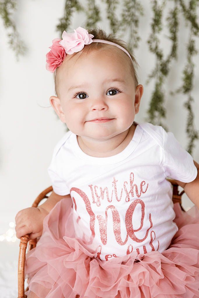 Baby girl in pink tutu and floral headband wearing an Isn't She One onesie sits smiling in a wicker chair.